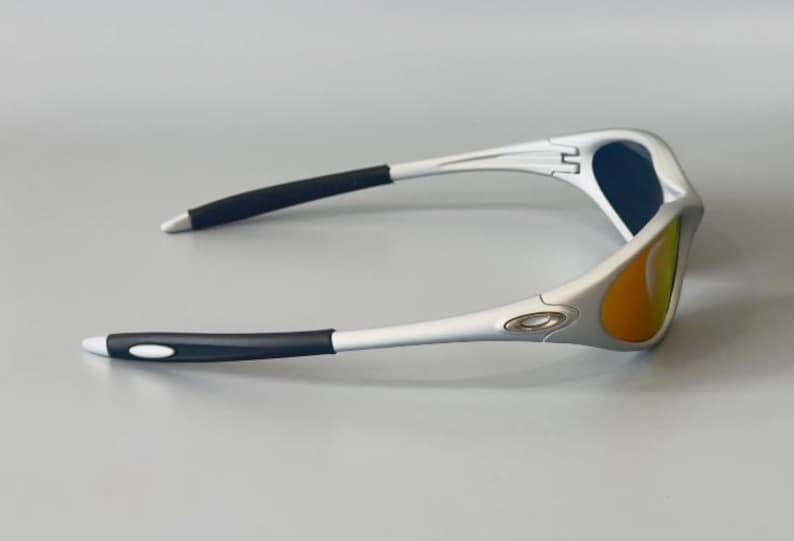 K&ouml;nnte beinhalten: Silberne Sonnenbrille mit schwarzen B&uuml;geln und blau-orangen Gl&auml;sern. Die Sonnenbrille hat ein sportliches Design. Das Oakley-Logo ist auf dem Rahmen sichtbar. Geeignet f&uuml;r Outdoor-Aktivit&auml;ten.