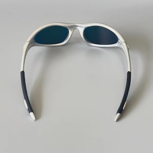K&ouml;nnte beinhalten: Silberfarbene Sonnenbrille mit dunklen Gl&auml;sern. Die Sonnenbrille hat ein schlankes, modernes Design mit wei&szlig;en B&uuml;geln und schwarzen Gummi-Griffen an den Enden. Die Gl&auml;ser sind dunkel und reflektierend.