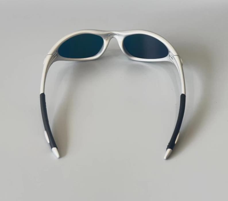K&ouml;nnte beinhalten: Silberfarbene Sonnenbrille mit dunklen Gl&auml;sern. Die Sonnenbrille hat ein schlankes, modernes Design mit wei&szlig;en B&uuml;geln und schwarzen Gummi-Griffen an den Enden. Die Gl&auml;ser sind dunkel und reflektierend.