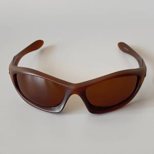May include: Brown sunglasses with a curved frame and tinted lenses. The sunglasses have a sporty design and are suitable for outdoor activities. The frame is a solid brown color.