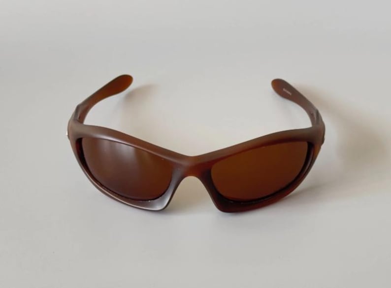 May include: Brown sunglasses with a curved frame and tinted lenses. The sunglasses have a sporty design and are suitable for outdoor activities. The frame is a solid brown color.