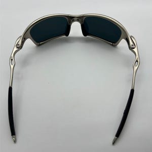 Puede incluir: Gafas de sol con montura plateada y lentes oscuras. Las gafas tienen un dise&ntilde;o curvado con agarres de goma negra en las patillas para mayor comodidad. Las lentes son de un color oscuro, proporcionando un aspecto elegante y moderno.