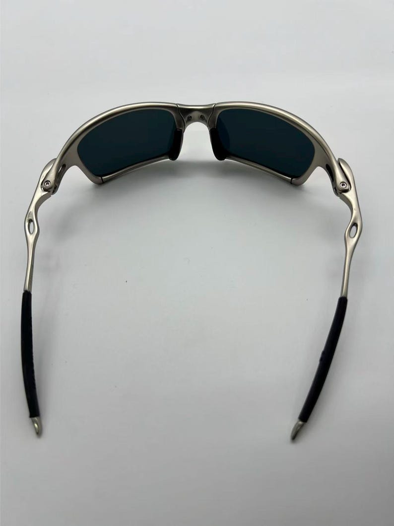 Puede incluir: Gafas de sol con montura plateada y lentes oscuras. Las gafas tienen un dise&ntilde;o curvado con agarres de goma negra en las patillas para mayor comodidad. Las lentes son de un color oscuro, proporcionando un aspecto elegante y moderno.