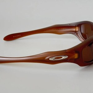 May include: Brown sunglasses with a curved frame and tinted lenses. The sunglasses have a silver emblem on the temples. The arms of the sunglasses are long and slender, with a slight curve.