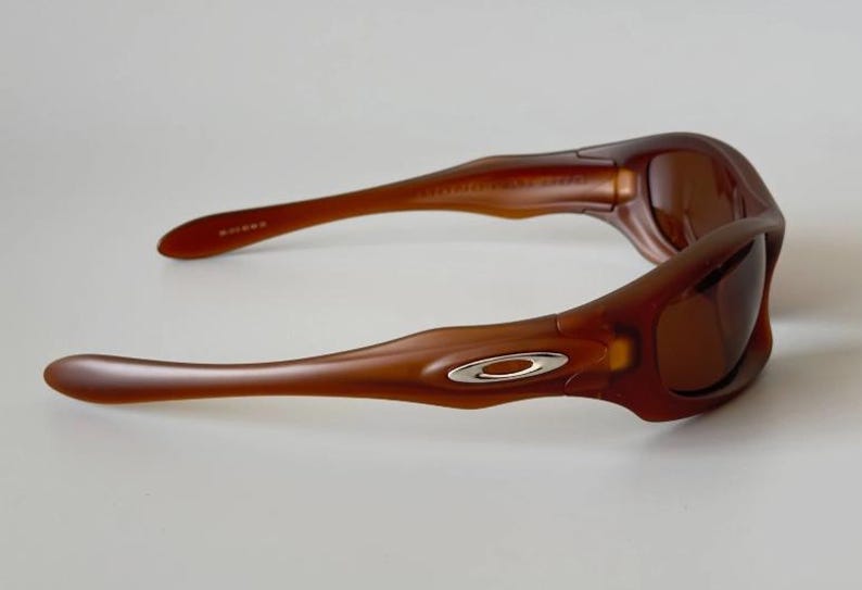 May include: Brown sunglasses with a curved frame and tinted lenses. The sunglasses have a silver emblem on the temples. The arms of the sunglasses are long and slender, with a slight curve.