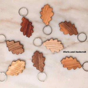 Könnte beinhalten: Eine Sammlung von Eichenblatt-Schlüsselanhängern aus Holz in verschiedenen Braun- und Beigetönen. Jeder Schlüsselanhänger hat einen silberfarbenen Metallring. Die Schlüsselanhänger liegen auf einer hellen Oberfläche.