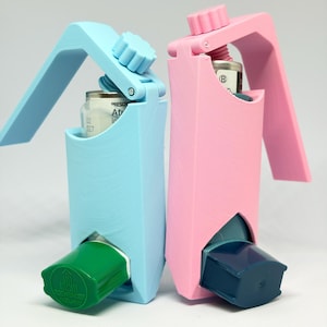 May include: Two 3D-printed inhaler holders, one light blue and one light pink, each featuring a top-mounted adjustment knob. The blue holder contains an inhaler with a green cap, while the pink holder contains an inhaler with a dark blue cap.