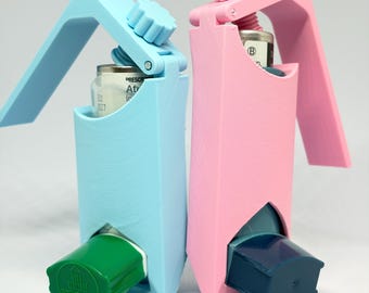 May include: Two 3D-printed inhaler holders, one light blue and one light pink, each featuring a top-mounted adjustment knob. The blue holder contains an inhaler with a green cap, while the pink holder contains an inhaler with a dark blue cap.
