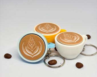 May include: Three miniature coffee cup keychains in light blue, yellow, and white, each featuring a detailed latte art design on the surface. The keychains are accompanied by several scattered dark brown coffee beans on a plain white background.