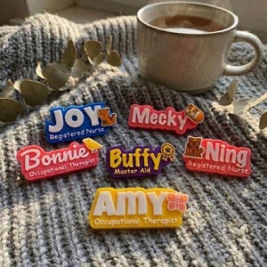Puede incluir: Una colección de coloridas placas con nombres como "Joy", "Mecky", "Bonnie", "Buffy", "Ning" y "Amy". Cada placa tiene un diseño único e incluye una profesión, como "Enfermera Registrada" o "Terapeuta Ocupacional". Una taza de té se encuentra sobre una superficie de punto gris.