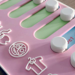 May include: A pink plastic activity board featuring white sliding knobs and white raised icons including a tooth, a pizza slice, and a shirt. The board has green and blue recessed tracks, with visible text reading "pet dresse" and "Mor" on the surface.