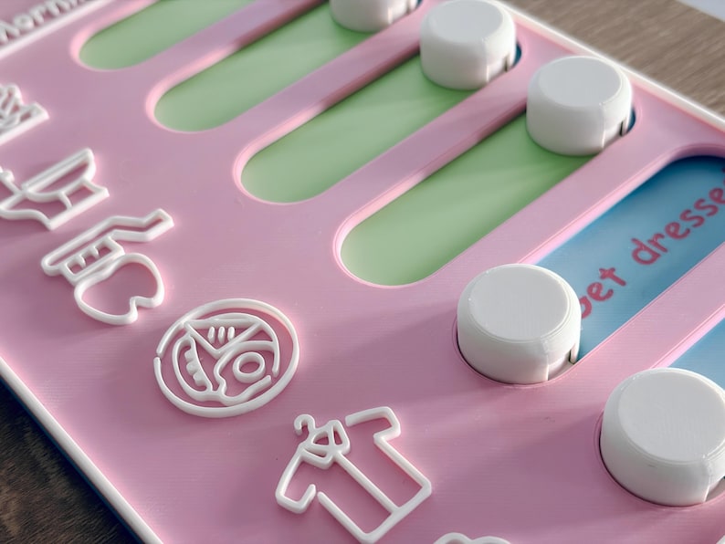 May include: A pink plastic activity board featuring white sliding knobs and white raised icons including a tooth, a pizza slice, and a shirt. The board has green and blue recessed tracks, with visible text reading "pet dresse" and "Mor" on the surface.