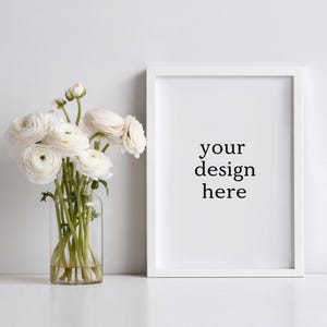 May include: A white picture frame with the text "your design here" stands next to a clear glass vase filled with white flowers. The flowers have multiple layers of petals.