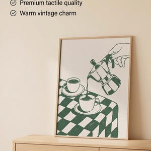 Könnte beinhalten: Ein gerahmter Wandkunstdruck mit einer grün-weißen Illustration einer Mokkakanne, die Kaffee in eine Tasse auf einer karierten Tischdecke gießt. Der Text lautet: The only wall art made for: Curated, calm spaces, Fade-resistant giclée inks, Premium tactile quality, Warm vintage charm, Atelier Maxi.