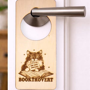 May include: Wooden door hanger featuring a cat illustration. The cat is reading a book and holding a sign that says "DO NOT DISTURB." The word "BOOKTROVERT" is below the book. The hanger is attached to a door handle.