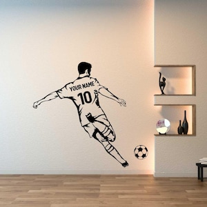 May include: Black and white wall decal of a soccer player kicking a soccer ball. The player is wearing a jersey with the number 10 and the text "YOUR NAME" on the back. The decal is on a white wall.