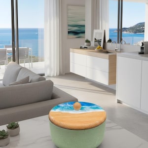 May include: A round, green container with a wooden lid featuring a beach scene design. The lid has a light brown wooden base with a blue and white wave design. A small, round wooden knob sits atop the lid. The container sits on a white marble table.