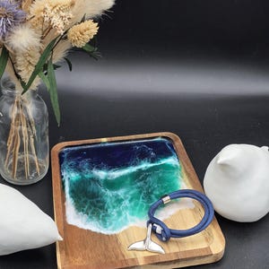 May include: A wooden tray with a resin ocean design in shades of blue and white. A blue rope bracelet with a silver whale tail charm rests on the tray. Decorative elements include a vase of dried flowers and a white ceramic bird.