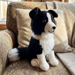 Könnte beinhalten: Ein handgehäkelter Hund im Border Collie-Stil in Schwarz und Weiß. Das Stofftier sitzt auf einem gemusterten Sessel, mit flauschiger weißer Brust, Pfoten und Schwanzspitze. Der Hund hat braune Augen und eine schwarze Nase.