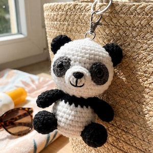 Puede incluir: Un llavero de panda tejido a mano en crochet, en blanco y negro. El panda tiene orejas, ojos, brazos y piernas negros, con cabeza y cuerpo blancos. Está sujeto a un cierre y un anillo de metal plateado. El llavero está junto a un bolso de paja trenzada.