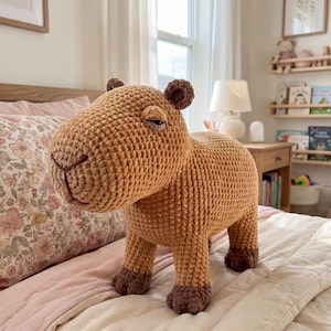 Op de afbeelding: Een groot, gehaakt capybaraknuffel in tinten bruin en beige. Het handgemaakte dier heeft een gedetailleerd, getextureerd oppervlak, kleine oren en donkerbruine voeten. Het staat op een bed met roze beddengoed, wat wijst op een kinderkamer.