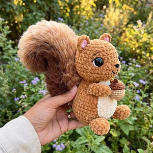 Op de afbeelding: Een gehaakte eekhoorn knuffel met een pluizige bruine en bruine staart. De eekhoorn heeft een bruin lichaam, een witte buik en houdt een kleine eikel vast. De eekhoorn heeft zwarte ogen en roze oren.