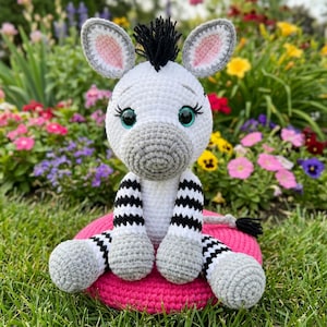 Może przedstawiać: Ręcznie robiona szydełkowa lalka zebry z białym korpusem, czarno-białymi paskami na nogach i ramionach oraz czarną grzywą. Zebra ma szare uszy i pysk i siedzi na różowej poduszce. Tło przedstawia kolorowe kwiaty i zieloną trawę.