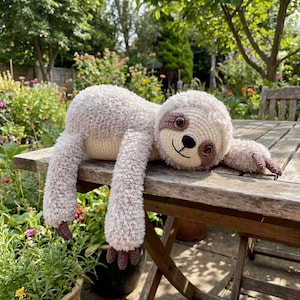 Könnte beinhalten: Ein handgefertigtes, plüschiges Faultier-Spielzeug in Beige- und Brauntönen, das auf einem Holztisch liegt. Das Faultier hat einen runden Kopf, lange Arme und kleine Füße. Sein Gesicht hat braune Augen und einen lächelnden Mund. Der Hintergrund ist grün.