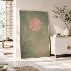 Puede incluir: Una impresión artística enmarcada que presenta un sol rosa y una bandada de pájaros sobre un fondo verde apagado. Una rama de árbol es visible en la esquina inferior izquierda. La obra tiene un marco dorado y está apoyada contra una pared blanca.