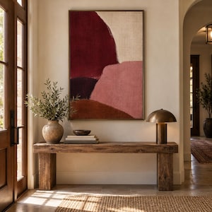 May include: A rectangular framed wall art piece featuring abstract color blocks in deep burgundy, muted pink, dark brown, and cream. The artwork hangs above a rustic wooden console table holding a ceramic vase with greenery, books, and a lamp.