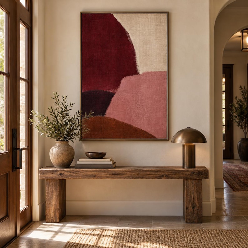 May include: A rectangular framed wall art piece featuring abstract color blocks in deep burgundy, muted pink, dark brown, and cream. The artwork hangs above a rustic wooden console table holding a ceramic vase with greenery, books, and a lamp.