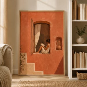 May include: A framed wall art print featuring a terracotta-colored wall with a person in a white dress sitting in an arched window reading a book. The scene includes a white curtain, a small arched niche with a vase, and light-colored stone steps.