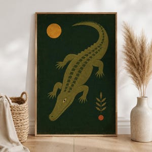 Könnte beinhalten: Ein rechteckiger Wandkunstdruck in einem Holzrahmen mit dunkelgrünem Hintergrund, einem großen olivgrünen Krokodil, einem goldgelben Kreis als Sonne, einem kleinen orangefarbenen Kreis und einer stilisierten grünen Blattzweig-Illustration.