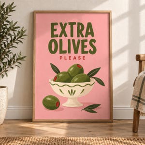 May include: A framed wall art print with a pink background featuring the text EXTRA OLIVES PLEASE in dark green and red. The illustration shows a white pedestal bowl filled with green olives and leaves, with one additional olive below.