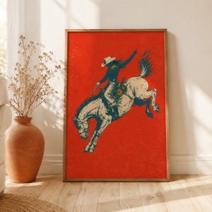 May include: A rectangular wall art print featuring a dark blue illustration of a person wearing a cowboy hat riding a bucking horse against a solid, textured red background. The artwork is framed in a simple, light-toned wooden frame.