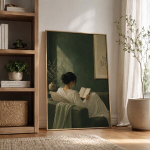 May include: A framed wall art print featuring a painting of a person with dark hair in a white dress sitting on a dark green sofa, reading an open book. The scene is set against a deep green wall with a light-coloured rectangular panel on the right.