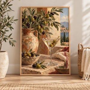 Può includere: Una stampa d'arte incorniciata che raffigura una scena mediterranea con un grande vaso in terracotta, una ciotola di olive verdi e nere e un bicchiere da martini contenente olive. Lo sfondo mostra un paesaggio costiero con un edificio in pietra.