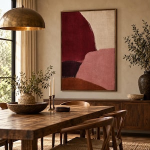 May include: A large rectangular wall art piece featuring abstract color blocks in deep burgundy, muted pink, cream, and earthy brown tones, framed in a thin wood border, displayed on a neutral wall above a wooden dining table and sideboard.