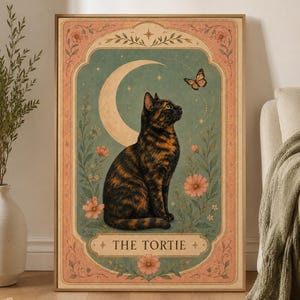 Könnte beinhalten: Ein rechteckiger Wandkunstdruck, der eine Schildpattkatze im Profil zeigt, die zu einem Schmetterling aufschaut. Der Hintergrund ist ein blaugrünes Feld mit einer Mondsichel, Blumen und Laub, umrahmt von einem rosa Rand mit dem Text THE TORTIE.