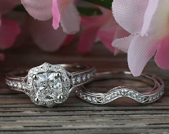 May include: A diamond engagement ring with a square-cut center stone and a matching curved wedding band. The rings are set with smaller diamonds and are displayed on a wooden surface, with pink flowers in the background.