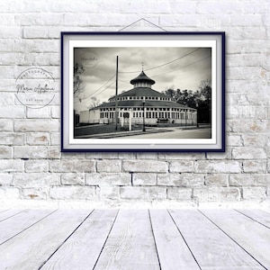 May include: Black and white framed photograph of a round building with a domed roof, likely a carousel or bandstand. The photograph is displayed on a white brick wall with a wooden floor. The photographer's name and establishment date are visible.