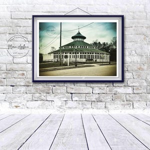May include: Framed print of a vintage carousel building with a green roof and white exterior. The image is in sepia tones, with a dark blue frame. The print is hanging on a white brick wall above a white wooden floor.