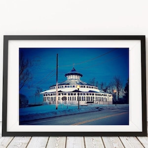 May include: A framed photograph of a round building with a domed roof, lit up at night. The building has many windows and is surrounded by snow. The sky is a deep blue, and the image is framed in black.