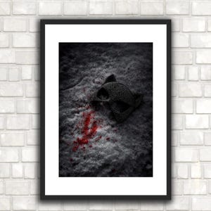 May include: A framed print featuring a black and silver mask resting on a textured, gray surface with red splatters. The mask has a detailed, patterned design. The image is set against a white brick wall.