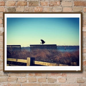 May include: Framed landscape art print showing a bird in flight over water. The image includes a wooden fence, benches, and tall grass. The sky is a gradient of blue and the frame is brown.