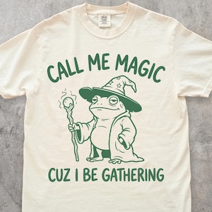 May include: Ivory t-shirt featuring a green frog wizard graphic with the text "CALL ME MAGIC CUZ I BE GATHERING". The frog wears a hat and robe, holding a staff. The shirt has a crew neck and short sleeves. The colour is ivory.