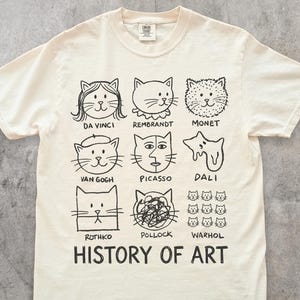 以下が含まれることがあります： アイボリー色のTシャツ。ダ・ヴィンチやウォーホルなど、有名なアーティストにインスパイアされた猫のポートレートの白黒イラストが描かれています。「HISTORY OF ART」の文字が猫のポートレートの下にプリントされています。