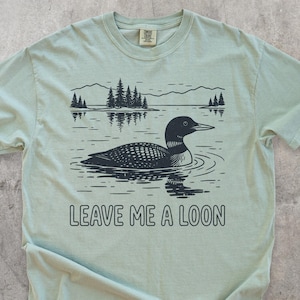 May include: A pale green t-shirt with a black illustration of a loon on a lake, with mountains and trees in the background. The text "LEAVE ME A LOON" is printed below the image. The shirt has a crew neck and short sleeves.