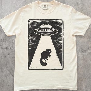 May include: Ivory t-shirt with a black print of a UFO abducting a cat. The design is in a rectangular frame. The shirt has a crew neck and short sleeves. The brand logo is visible on the upper right.