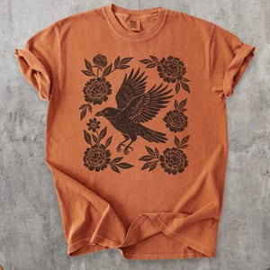 May include: Burnt orange t-shirt featuring a black raven with outstretched wings, surrounded by floral elements. The design is centered on the shirt, with rolled sleeves. The shirt has a vintage, slightly faded appearance.